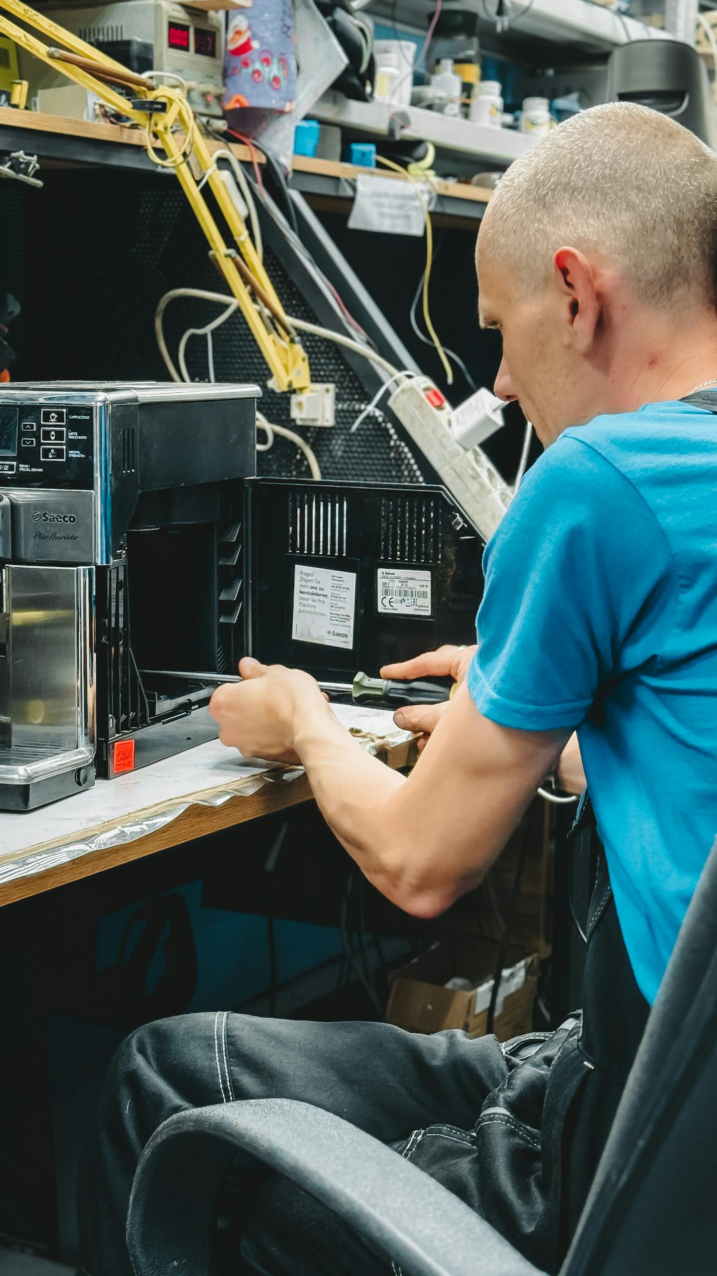 A skilled technician repairs a coffee machine in a cluttered electronic workshop.