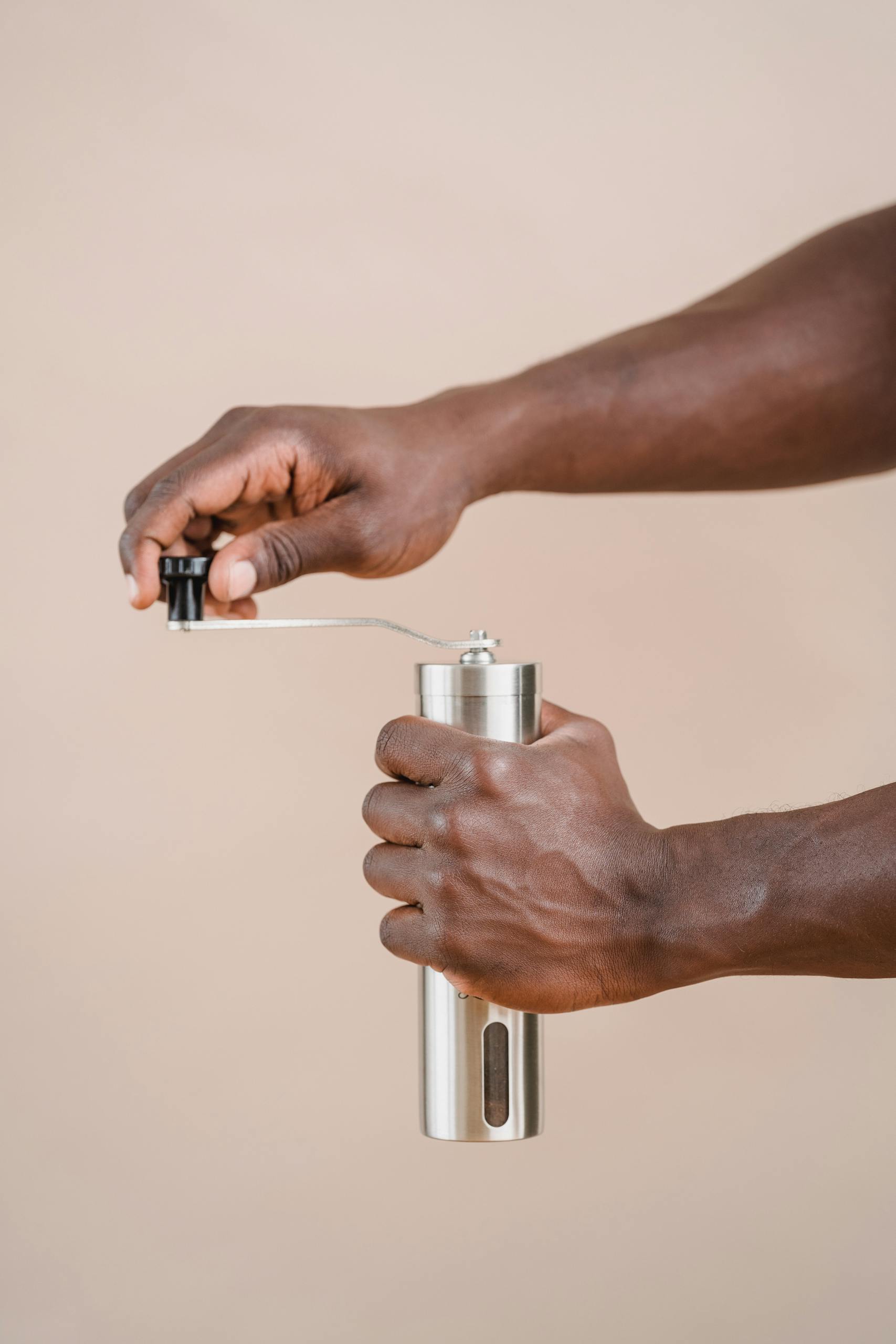A close-up image of a person using a stainless steel grinder with a beige background.
