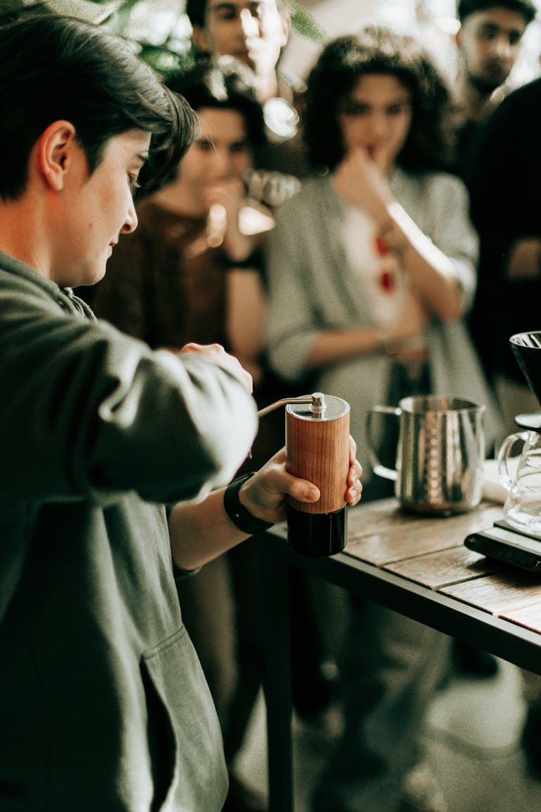 A barista demonstrates coffee grinding technique in a café with an attentive audience watching.