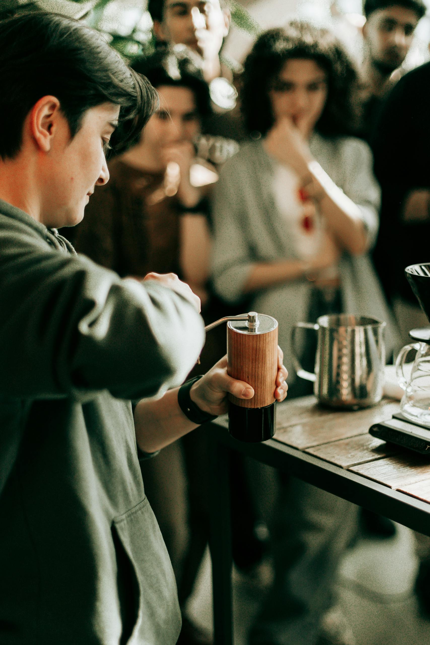 A barista demonstrates coffee grinding technique in a café with an attentive audience watching.