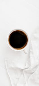 A minimalist overhead shot of a black coffee in a white cup on a white surface with a textured white towel.