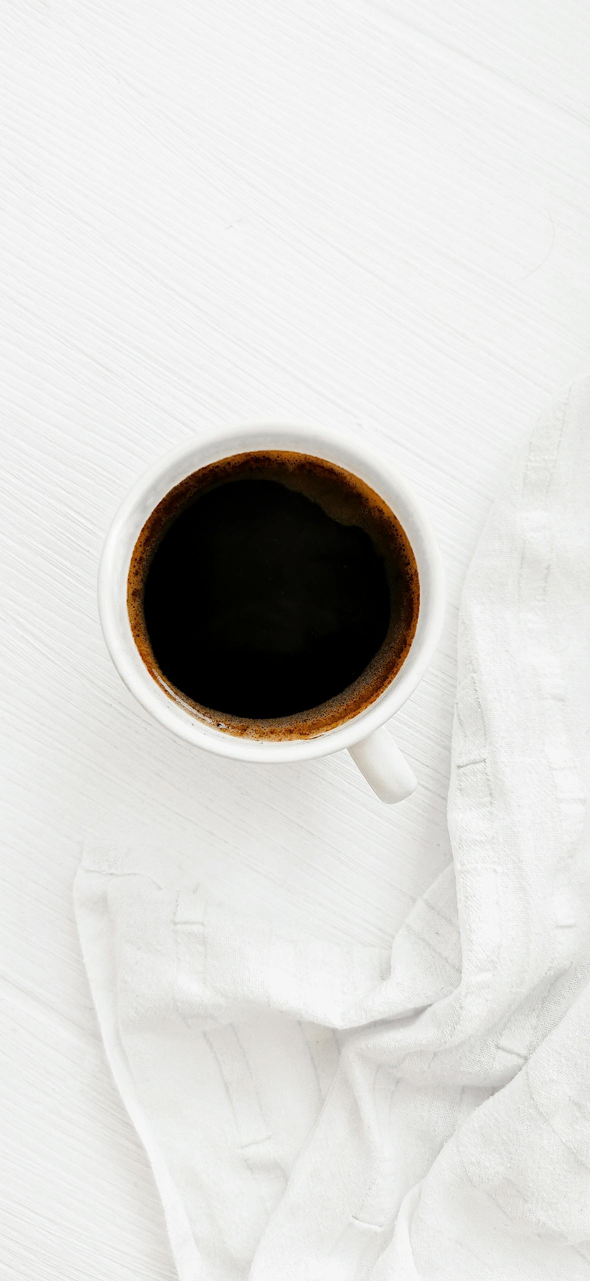 A minimalist overhead shot of a black coffee in a white cup on a white surface with a textured white towel.